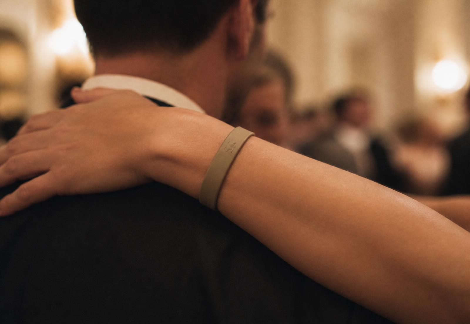 A dancer's wrist resting on a partner's shoulder, wearing a slim matte Sojourn band embossed with the S monogram